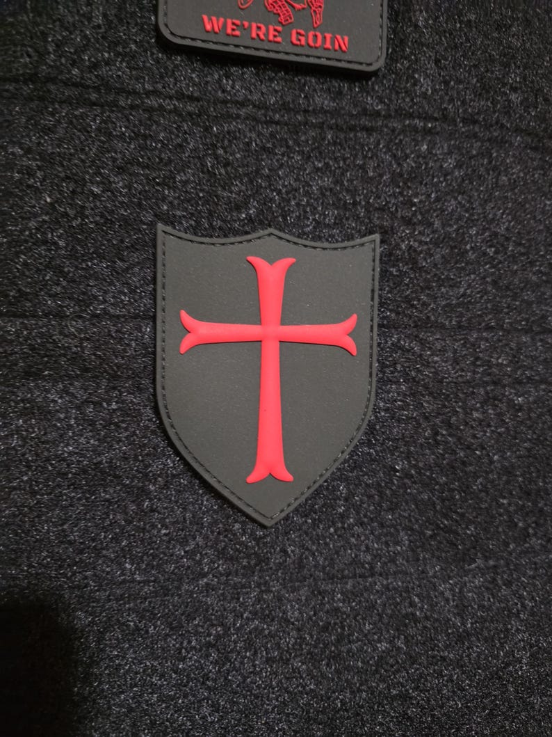 May include: Two patches are shown. The first is a black shield with a red cross. The second is a black rectangle with the words "WE'RE GOIN" in red. Both patches are on a dark gray textured background.