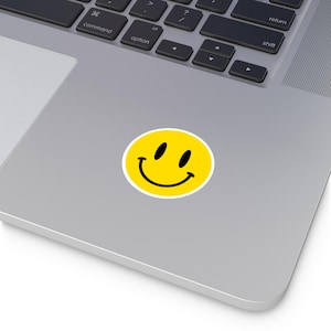 Puede incluir: Una pegatina de cara sonriente amarilla con un contorno blanco y ojos y boca negros. La pegatina está en un portátil plateado.