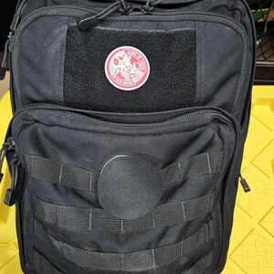 May include: Black tactical backpack with a red zipper pull and a red patch with the text "Hang in there it gets worse" and a graphic of a skull with a bandana.