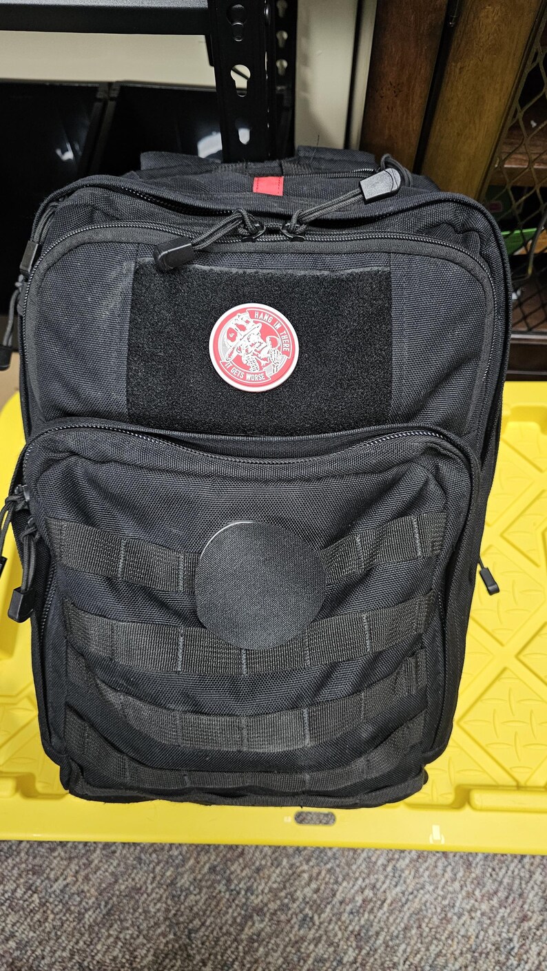 May include: Black tactical backpack with a red zipper pull and a red patch with the text "Hang in there it gets worse" and a graphic of a skull with a bandana.