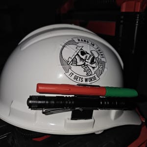 May include: White hard hat with a black and white sticker that says "Hang in there it gets worse" with a skull wearing a hard hat. A red and green marker and a black flashlight are attached to the hard hat.
