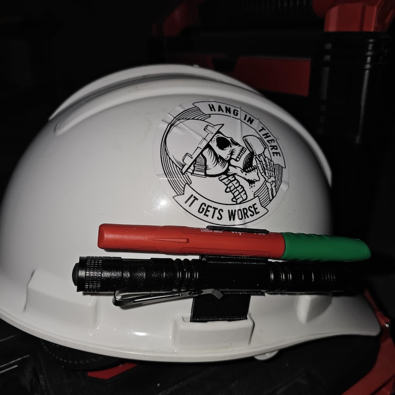 May include: White hard hat with a black and white sticker that says "Hang in there it gets worse" with a skull wearing a hard hat. A red and green marker and a black flashlight are attached to the hard hat.