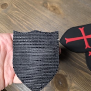 May include: A black shield-shaped patch with a textured surface. The patch is held in a hand, with other patches featuring a red cross design visible in the background. The patches appear to be made of fabric.