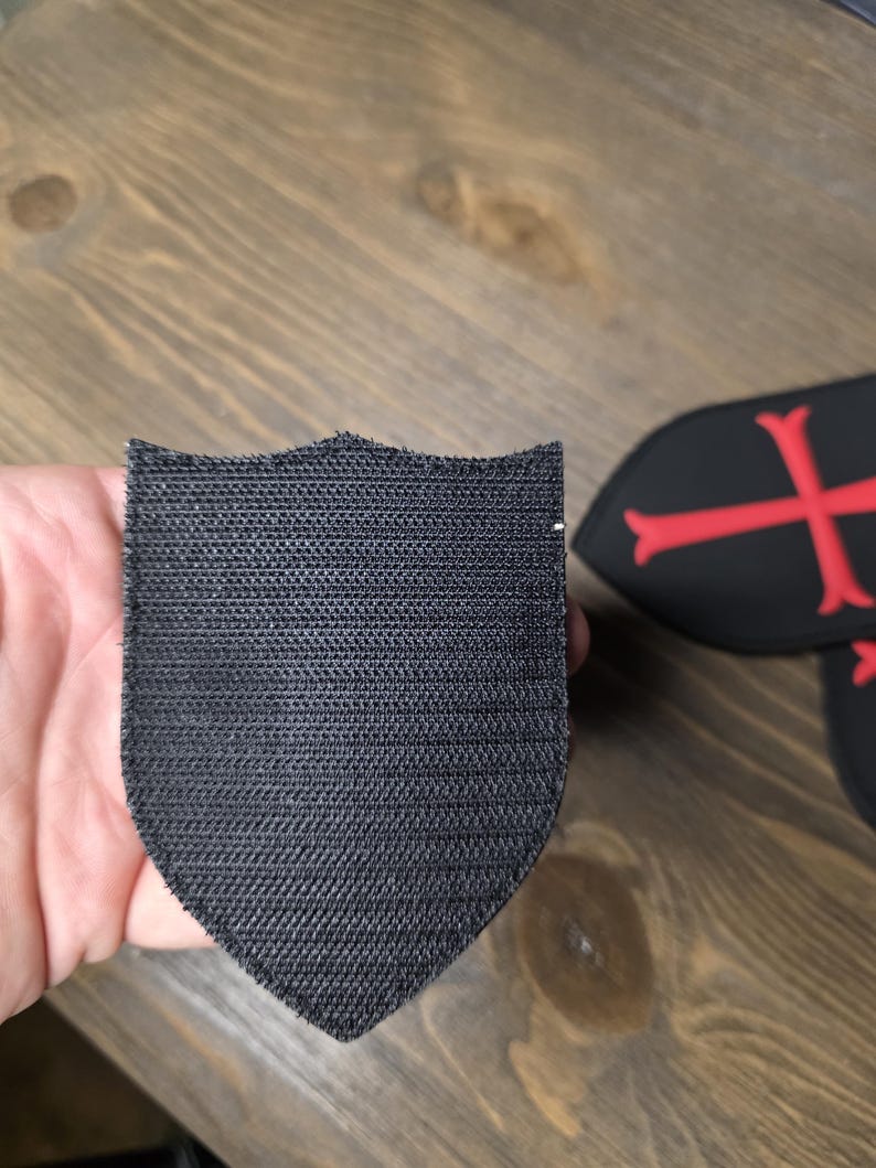 May include: A black shield-shaped patch with a textured surface. The patch is held in a hand, with other patches featuring a red cross design visible in the background. The patches appear to be made of fabric.