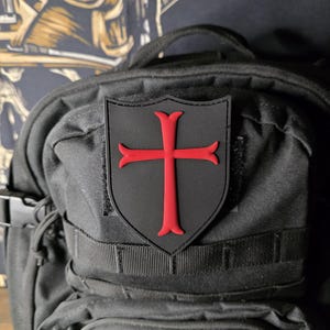 May include: A black shield-shaped patch with a red cross design, affixed to a black backpack. The cross has flared ends. The backpack has multiple straps and pockets.