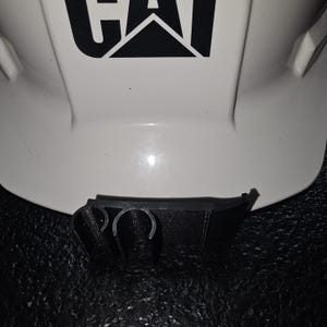 May include: A white hard hat with a black "CAT" logo and a black plastic clip attached to the bottom.