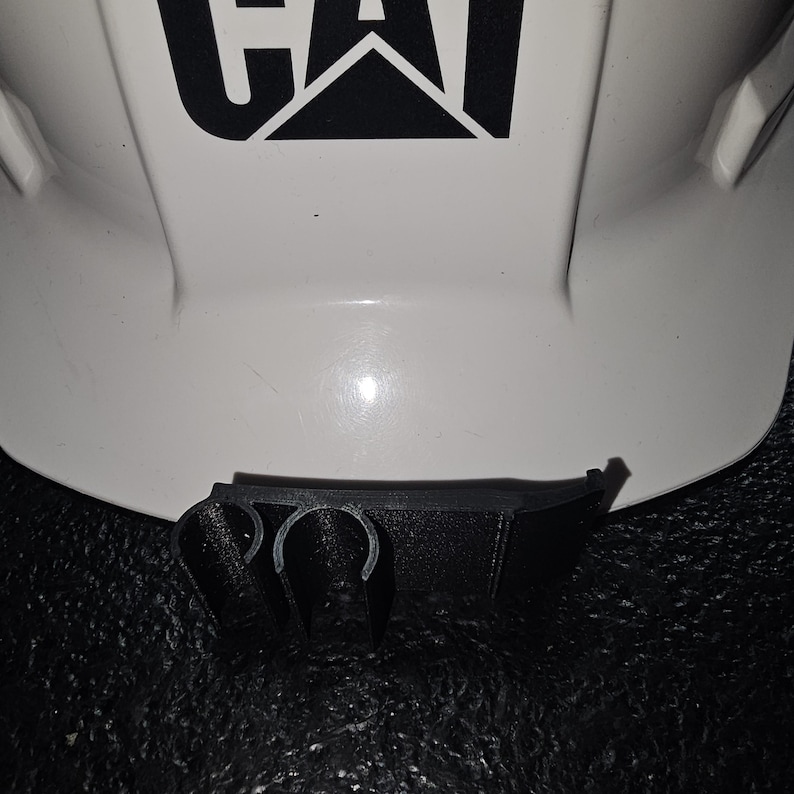 May include: A white hard hat with a black "CAT" logo and a black plastic clip attached to the bottom.