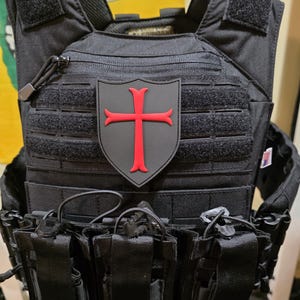 May include: Black tactical vest with multiple pouches and a red cross emblem. The vest has adjustable straps, a zip closure, and attachment points for extra gear. The emblem is a red cross on a grey background.