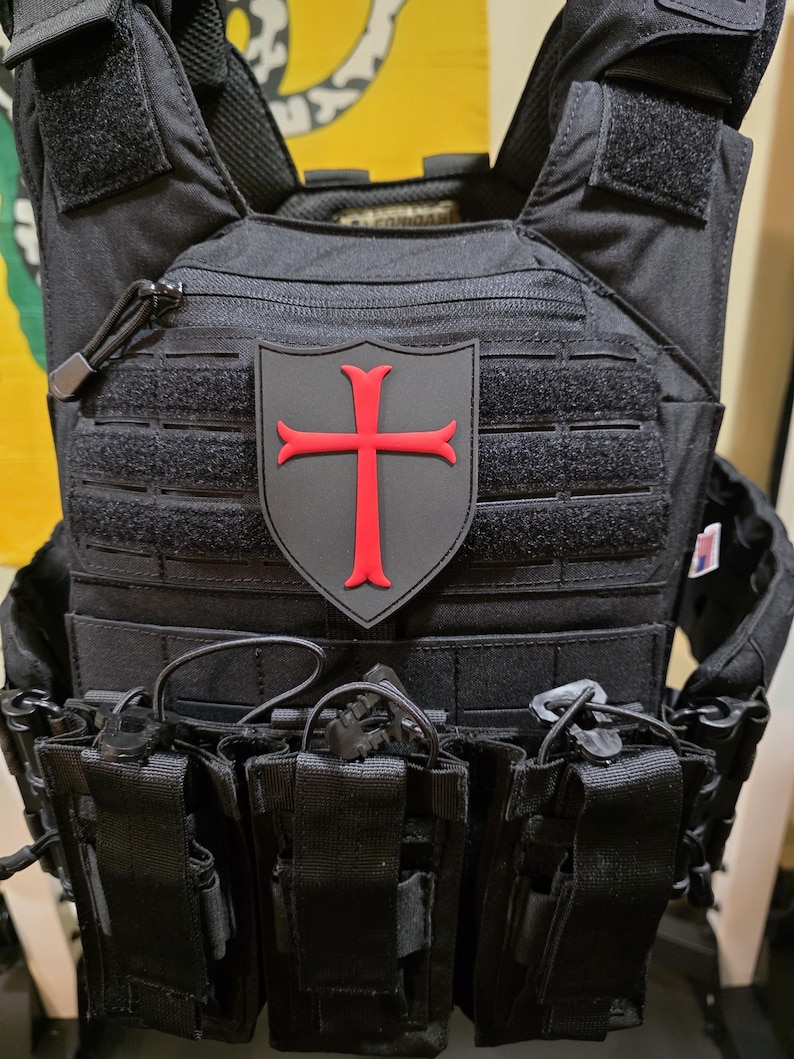 May include: Black tactical vest with multiple pouches and a red cross emblem. The vest features adjustable straps, a zipper closure, and various attachment points for additional gear. The emblem is a red cross on a gray background.