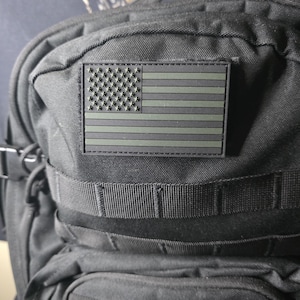 May include: Black tactical backpack with an olive green and black American flag patch. The backpack features multiple straps and compartments, and is made of a durable, textured material.