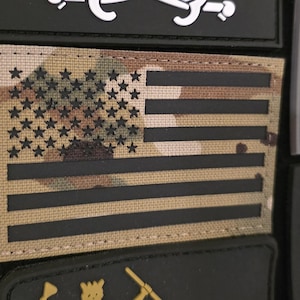 Könnte beinhalten: Eine Sammlung taktischer Aufnäher, darunter ein Tarnmuster-Aufnäher mit der amerikanischen Flagge mit schwarzen Sternen und Streifen. Andere Aufnäher zeigen militärische und patriotische Designs. Die Aufnäher sind an einem schwarzen Stoff befestigt.