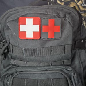 以下が含まれることがあります： 黒色のタクティカルバックパック。2つの正方形のパッチが付いています。1つは赤い縁と白い十字、もう1つは灰色の縁と赤い十字です。バックパックには複数のコンパートメントとストラップがあります。