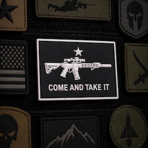Può includere: Collezione di toppe tattiche in varie forme e colori. Una toppa centrale presenta un design di fucile bianco con la scritta "COME AND TAKE IT". Altre toppe includono una stella, una bandiera americana, un teschio e una catena montuosa.