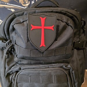 May include: Black backpack with multiple compartments and straps. A patch with a red cross design is attached to the front. The cross is a bold, vibrant red against a black background.