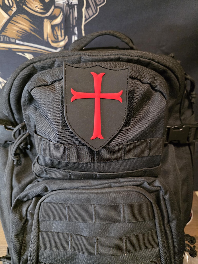 May include: Black backpack with multiple compartments and straps. A patch with a red cross design is attached to the front. The cross is a bold, vibrant red against a black background.