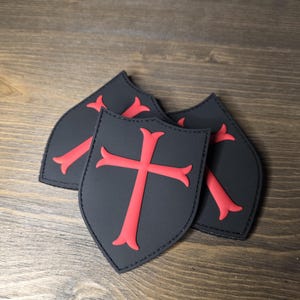May include: Three black shield-shaped patches with a red cross design. The patches are arranged on a wooden surface. The text "BUILT FOR" is visible in the background.