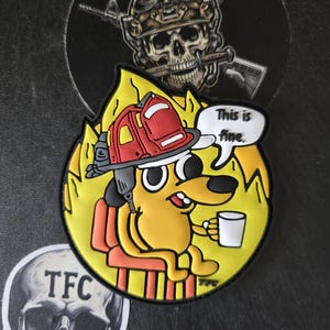 May include: A yellow patch featuring a cartoon dog in a firefighter's helmet, sitting in a burning room, holding a mug. A speech bubble says, "This is fine." Additional skull stickers are visible.