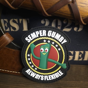 May include: A black patch with the words "Semper Gumby Always Flexible" encircling a green cartoon character with a red circle background. The patch has a yellow rope border and white stars.