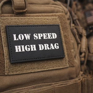 May include: Close-up of a tactical vest with a black patch. The patch reads "LOW SPEED HIGH DRAG" in white capital letters. The vest is tan and has various straps and attachments.