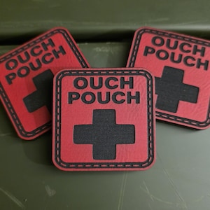 Pode incluir: Três patches quadrados vermelhos com as palavras "OUCH POUCH" e um símbolo de cruz negra. Os patches têm uma borda costurada e estão dispostos sobre uma superfície verde. O texto e a cruz estão em negrito.