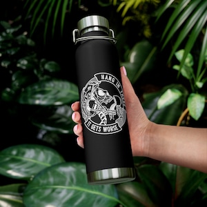 May include: Black water bottle with a silver lid and base. The bottle features a white graphic of a skull with the text "HANG IN THERE" and "IT GETS WORSE". The bottle is held against a backdrop of green leaves.