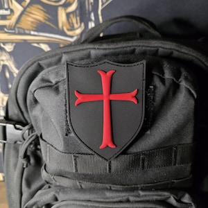 May include: A black shield-shaped patch with a red cross emblem, affixed to a black backpack. The cross has flared ends. The backpack has multiple straps and pockets, and a buckle closure.