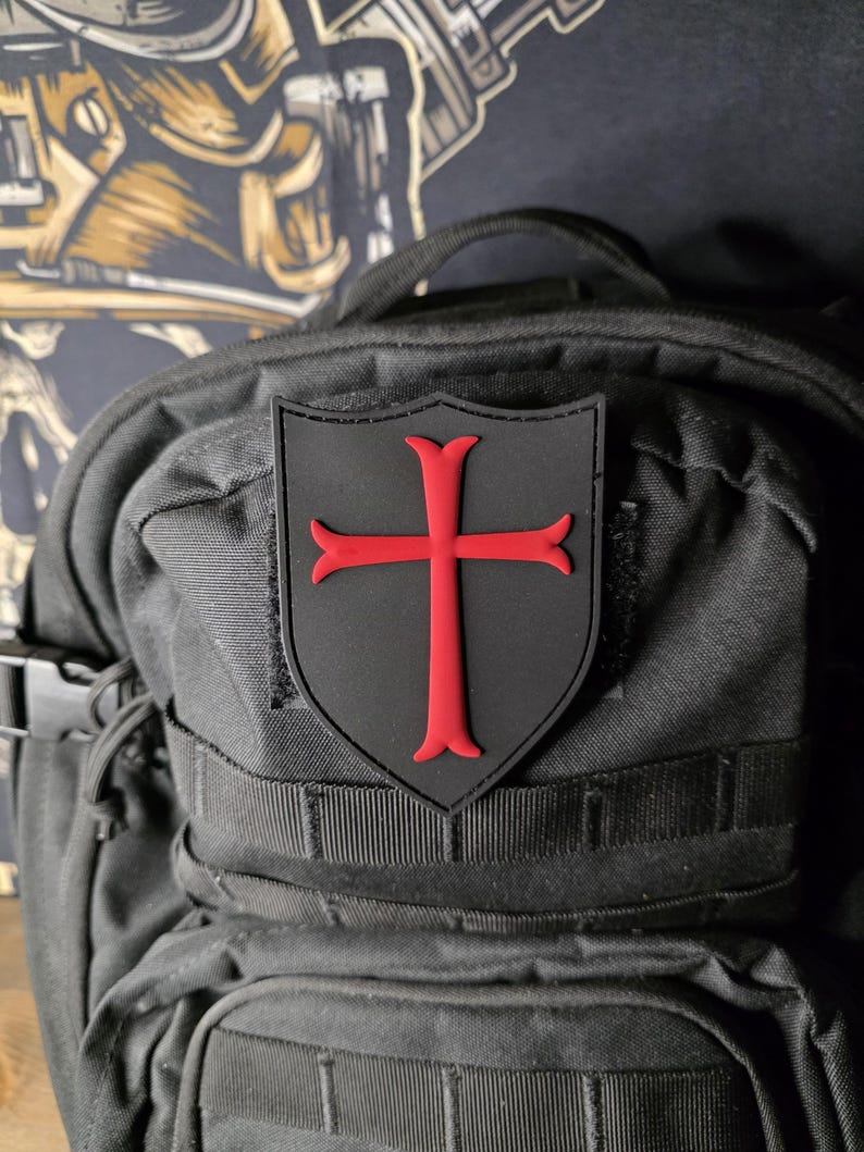 May include: A black shield-shaped patch with a red cross emblem, affixed to a black backpack. The cross has flared ends. The backpack has multiple straps and pockets, and a buckle closure.