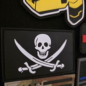 May include: A selection of patches, featuring a black rectangle with a white skull and crossed swords, a yellow and black design, and a camouflage American flag. The patches are displayed on a black surface.