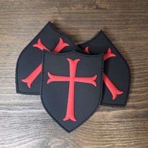 May include: Three black shield-shaped patches with a red cross design. The patches are arranged on a wooden surface. The crosses have a stylized, symmetrical design.