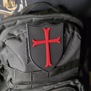 May include: A black patch with a red cross emblem, affixed to a black backpack. The cross is a bold, stylized design, set against a shield-shaped background. The patch is likely made of durable material.
