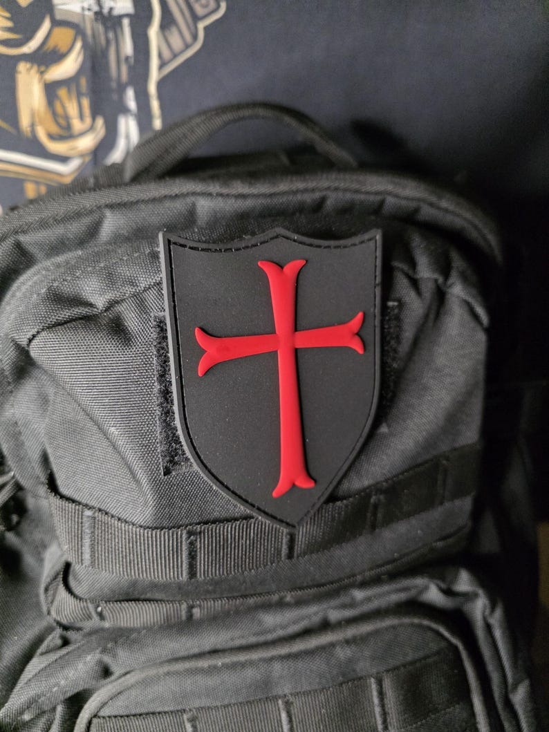 May include: A black patch with a red cross emblem, affixed to a black backpack. The cross is a bold, stylized design, set against a shield-shaped background. The patch is likely made of durable material.