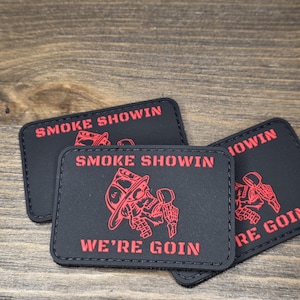 以下が含まれることがあります： 赤色の文字とグラフィックが入った、黒色の長方形のパッチが3つ。パッチには、消防士のヘルメットをかぶり、道具を持っている頭蓋骨が描かれています。パッチのテキストは「SMOKE SHOWIN」と「WE'RE GOIN」です。