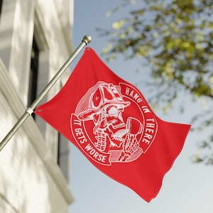 May include: A red flag with a white graphic of a firefighter skull wearing a helmet and holding a tool. The flag has the words "HANG IN THERE" and "IT GETS WORSE" in white text. The flag is mounted on a silver pole.