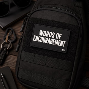 May include: A black tactical sling bag featuring a rectangular patch with the white text "WORDS OF ENCOURAGEMENT" and "TFC" in the corner. The bag includes a zippered compartment, a hook-and-loop panel, and horizontal webbing straps on the front.