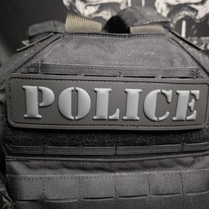 Puede incluir: Chaleco táctico negro con la palabra "POLICE" en letras grises en relieve. El chaleco tiene múltiples correas y un patrón de rejilla en la parte delantera. El fondo presenta un diseño de calavera.