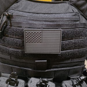 Peut inclure: Gilet tactique noir avec un écusson du drapeau américain. Le gilet comporte plusieurs poches et sangles, avec trois pochettes pour chargeurs visibles en bas. L'écusson du drapeau est de couleur sombre.