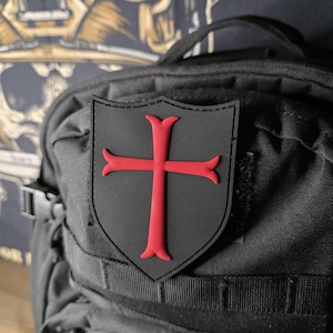 May include: Black shield-shaped patch with a red cross emblem, affixed to a black backpack. The patch is a prominent feature, contrasting against the textured fabric of the bag. The backpack has multiple compartments and straps.