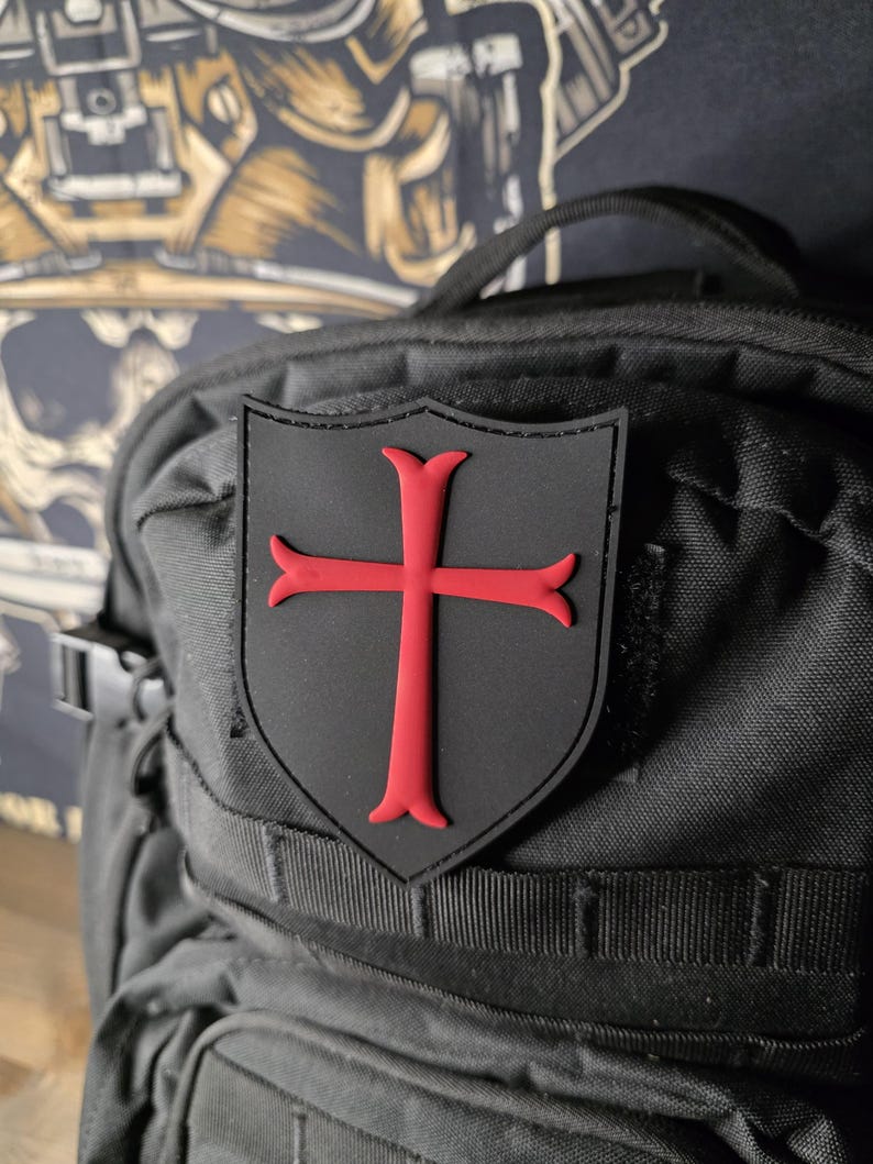 May include: Black shield-shaped patch with a red cross emblem, affixed to a black backpack. The patch is a prominent feature, contrasting against the textured fabric of the bag. The backpack has multiple compartments and straps.