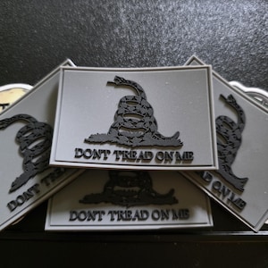Puede incluir: Varios parches grises con una serpiente de cascabel negra y el texto "DON'T TREAD ON ME". Los parches son rectangulares con un borde blanco. La imagen muestra un primer plano de los parches.