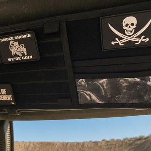 May include: A black vehicle sun visor organizer featuring MOLLE webbing, a clear plastic pocket, and several patches. The patches display a white cross, a skull and crossbones, and text reading SMOKE SHOWIN WE'RE GOIN and WORDS OF ENCOURAGEMENT.