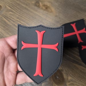 May include: Black shield-shaped patch with a red cross design. The patch has a stitched border and a textured surface. Additional patches are visible in the background. The patches are likely for clothing or accessories.