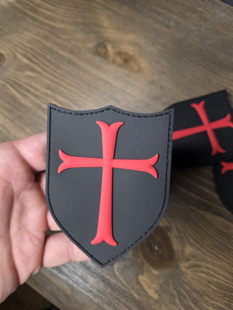 May include: Black shield-shaped patch with a red cross design. The patch has a stitched border and a textured surface. Additional patches are visible in the background. The patches are likely for clothing or accessories.