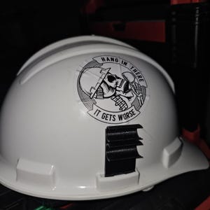 May include: White hard hat with a black and white sticker that says "Hang in there it gets worse" and a skull wearing a hard hat.