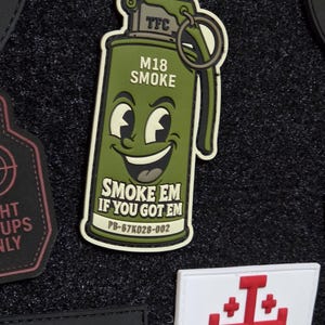 May include: An olive green rubberized morale patch shaped like a smoke grenade with a cartoon face. The text reads TFC, M18 SMOKE, SMOKE EM IF YOU GOT EM, and PB-67K028-002. The design features a pull ring, a lever, and a smiling character face.
