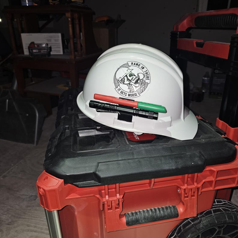 May include: A white hard hat with a black and white sticker that reads 'Hang in there it gets worse' and a skull and crossbones graphic. The hard hat has two black and green markers attached to it.
