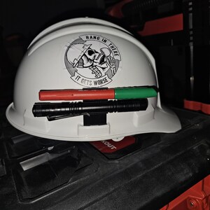 May include: A white hard hat with a black and white sticker that says "Hang in there, it gets worse". The sticker features a skull wearing a hard hat. A black flashlight and two red and green markers are attached to the hard hat.