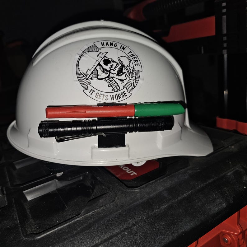 May include: A white hard hat with a black and white sticker that says "Hang in there, it gets worse". The sticker features a skull wearing a hard hat. A black flashlight and two red and green markers are attached to the hard hat.