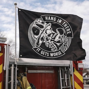 Puede incluir: Bandera negra con un gráfico blanco de una calavera con un casco de bombero, con el texto "HANG IN THERE IT GETS WORSE". La bandera está montada en un camión de bomberos rojo.