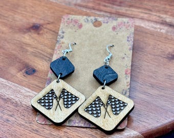 May include: Wooden earrings featuring a racing flag design. The earrings have a natural wood finish with black accents and silver-colored ear wires. The design includes two crossed checkered flags on a diamond-shaped pendant.