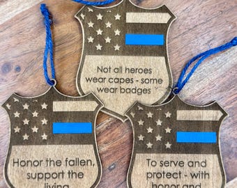 Könnte beinhalten: Drei schildförmige Holzornamente mit blauer Schnur. Jedes Schild hat eine blaue Linie, Sterne und Text: "Not all heroes wear capes - some wear badges", "Honor the fallen, support the living" und "To serve and protect - with honor and courage".